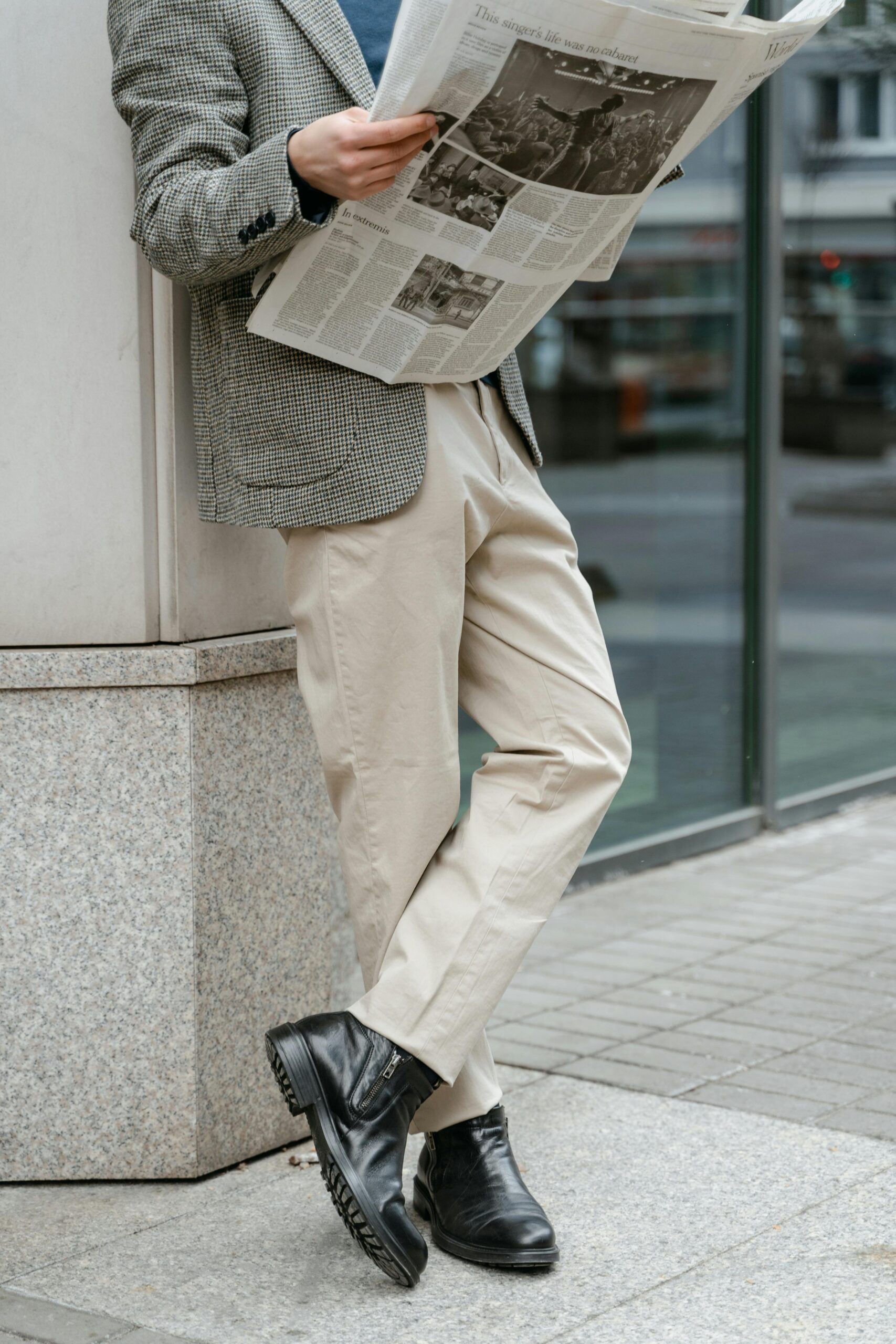 Man leaning against a wall reading a newspaper, wearing beige casual trousers, a patterned blazer, and black leather boots in a city setting.