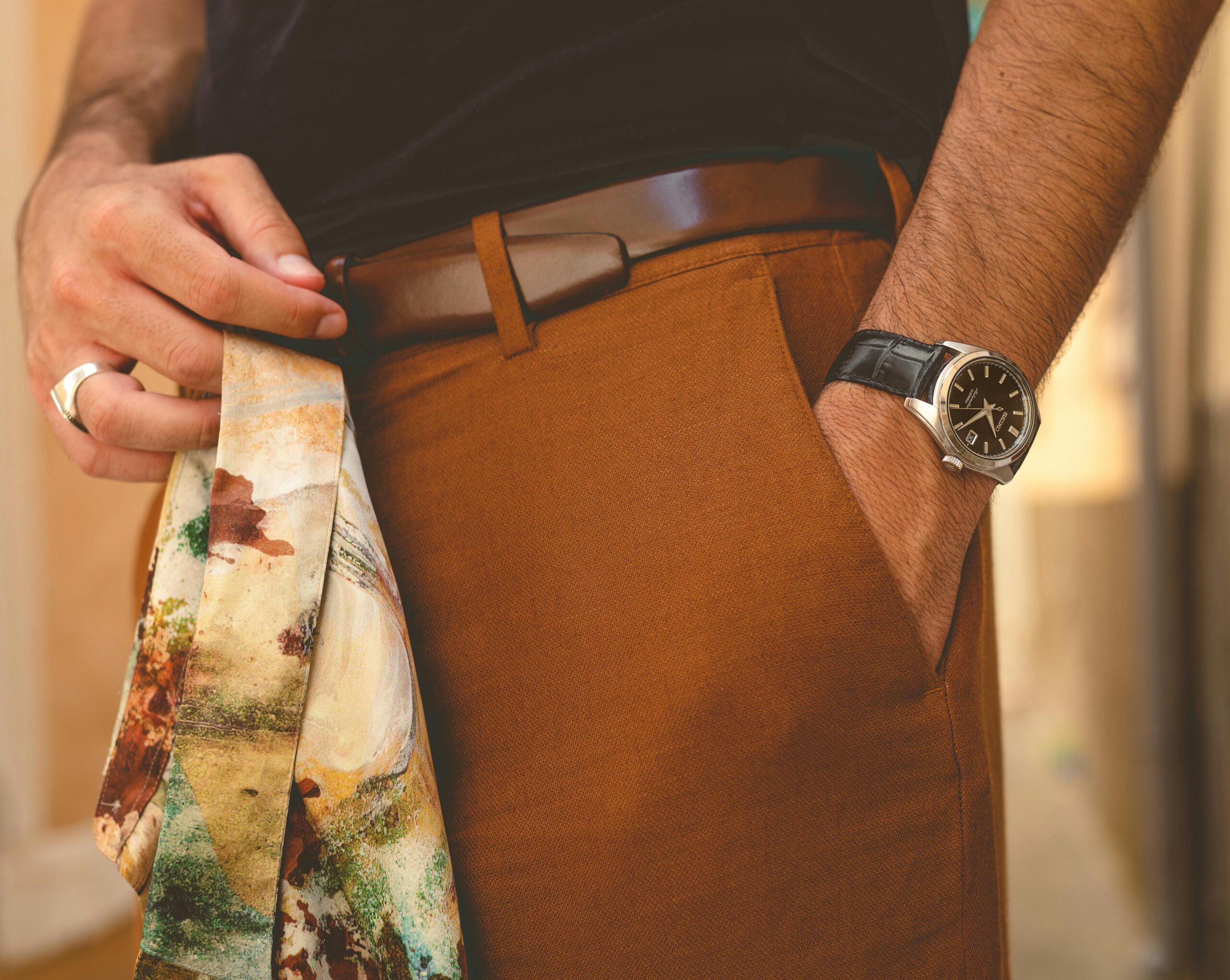 Close-up of brown men’s trousers with a leather belt, paired with a wristwatch and a patterned tie held in hand, highlighting smart-casual styling.